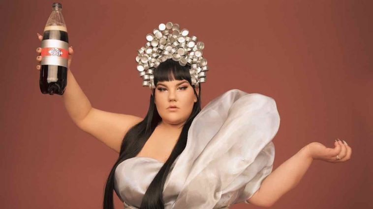 Netta wearing a silver crown and a silver dress with a big shoulder-padded strap, holding up what looks like a big bottle of Diet Coke but actually has the words "Ricki Lake" branded on it.