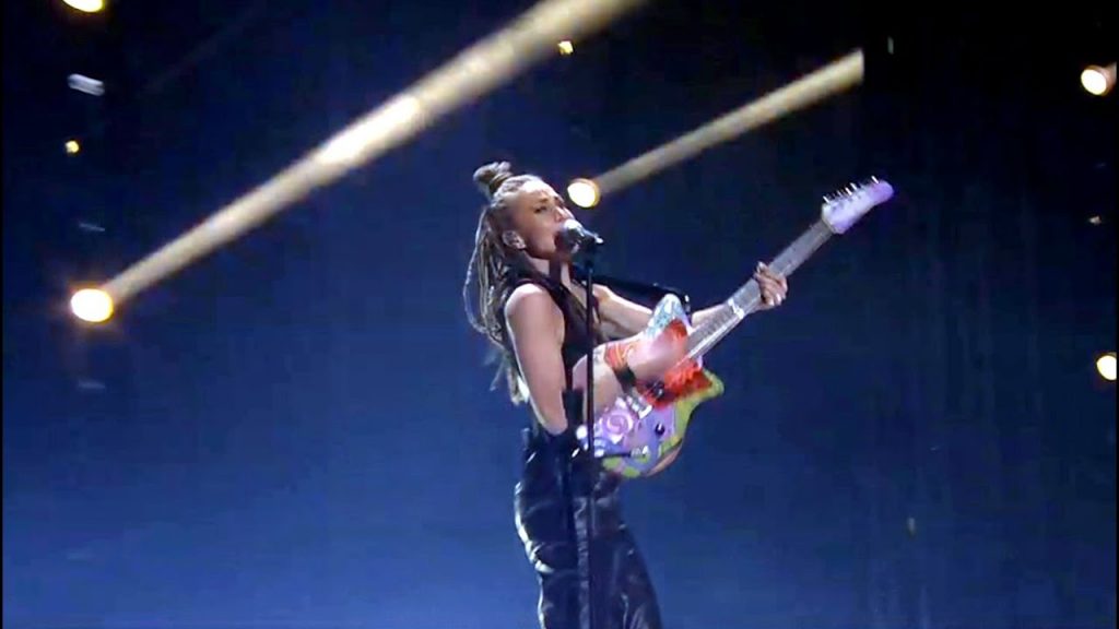 Mariette dressed in black with her hair in plats, playing a colourful guitar whilst singing her song "Shout It Out" on the Melodifestivalen 2020 stage.