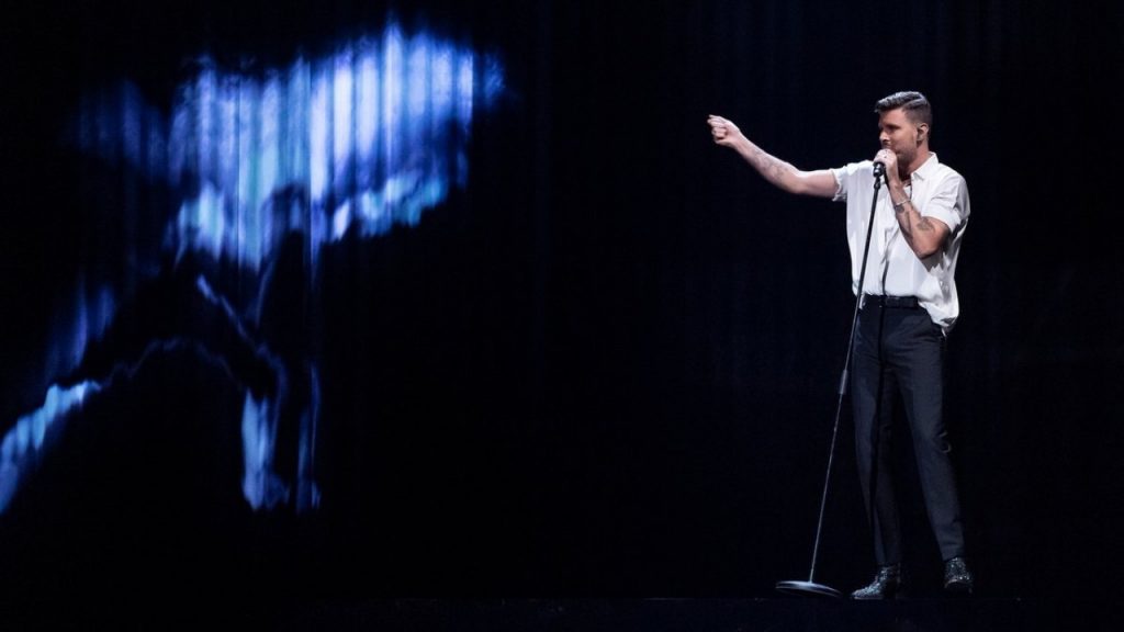 Robin Bengtsson competing in Melodifestivalen wearing a white shirt half-tucked into jeans as he points to a screen to his right which is displaying a figure in motion