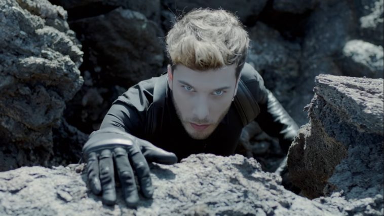 Blas Cantó "Universo" still which sees him climbing a rock ledge where the camera is, with a gloved hand on the top of the rock while he stares at the camera.