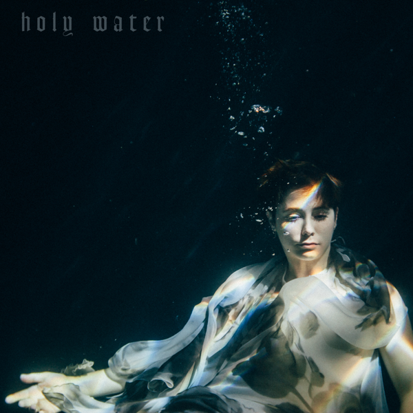 CeCe submerged in water wearing a flowy white top with the words "Holy Water" in the top left