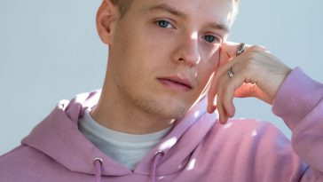Eurovision 2020 Germany representative Ben Dolic wearing a pink hoodie with his left hand leaning against his face