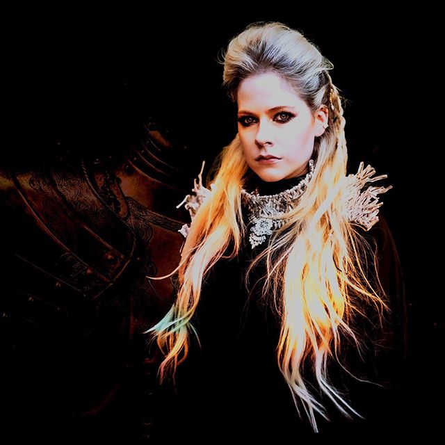 Avril Lavigne promo photo for her charity single "Warrior" wearing a silver neck armour piece with the background displaying a shadow of body armour.