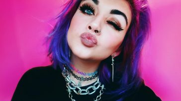 Bronnie promo pic for "Over You" which sees her winking at the camera, pouting, with her purple hair cut in a bob. She's wearing a black top and chain necklaces with a pink backdrop.