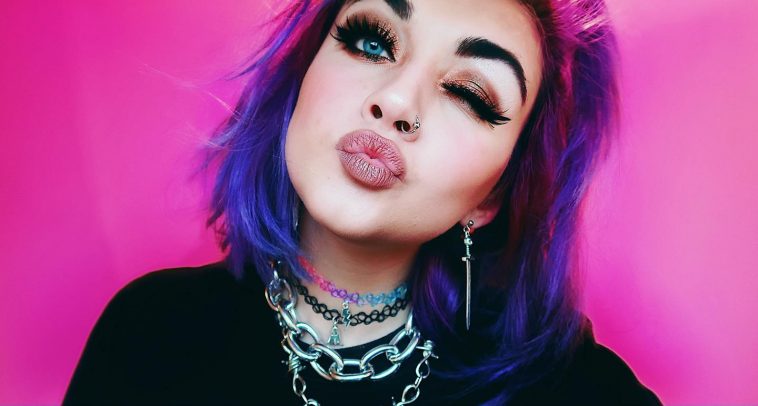 Bronnie promo pic for "Over You" which sees her winking at the camera, pouting, with her purple hair cut in a bob. She's wearing a black top and chain necklaces with a pink backdrop.