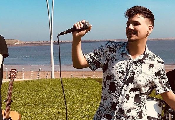 Danny Dearden performing in South Shields wearing a black and white shirt on some grass by the sea, with a microphone in his hand.
