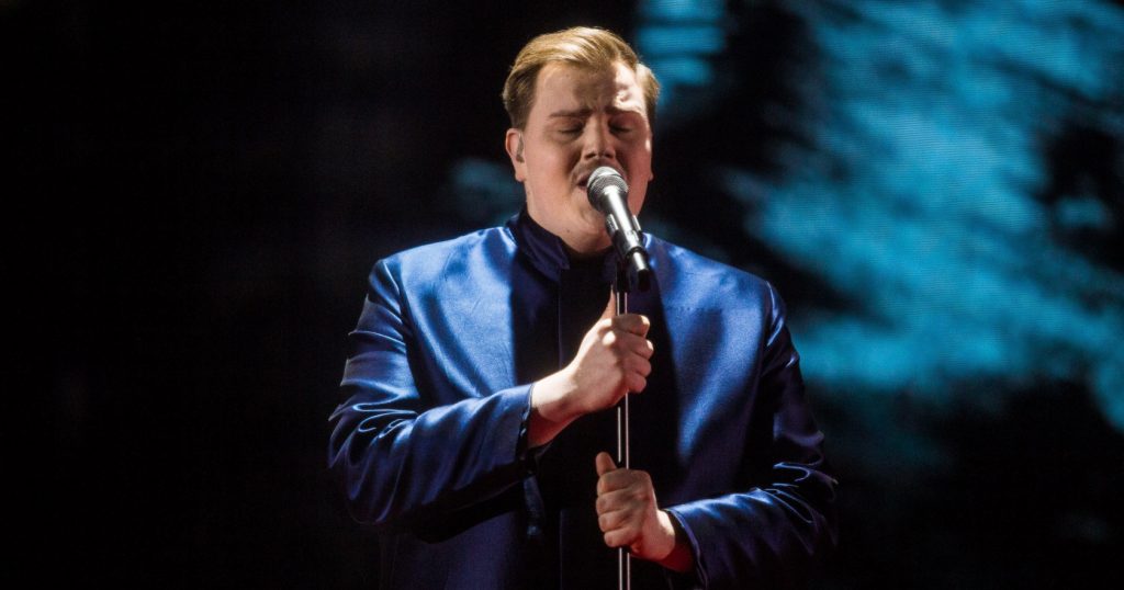 Aksel wearing a blue jacket over a black top, singing his song "Looking Back" on a stage at Finland's national selection show for Eurovision 2020.
