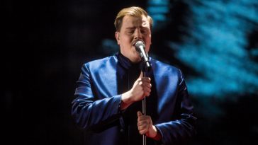 Aksel wearing a blue jacket over a black top, singing his song "Looking Back" on a stage at Finland's national selection show for Eurovision 2020.