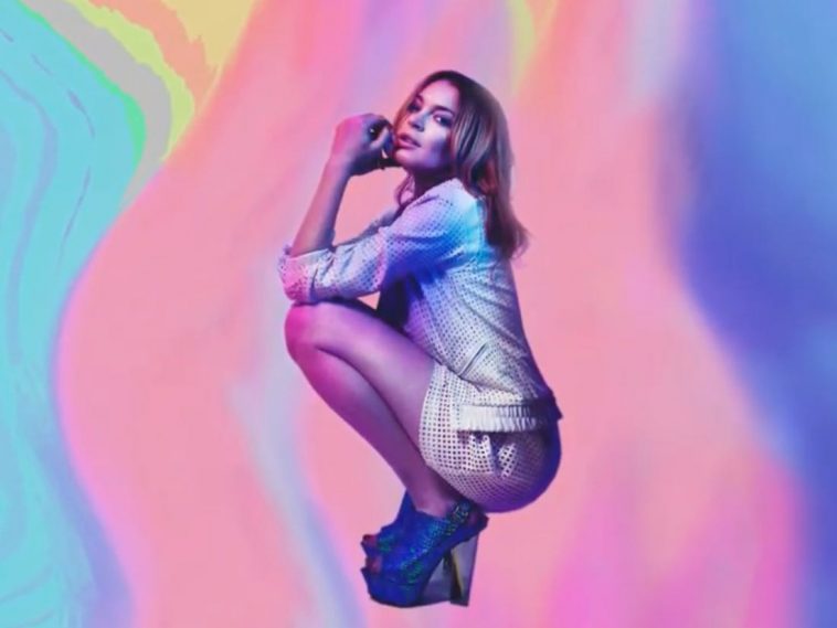 Lindsay Lohan posing for her "Back To Me" single artwork. She is crouching down with her elbows on her knees, she's wearing blue heels paired with a sky-blue top and gold short shorts backed by a blended paint streaks of light blue, pink, yellow and purple.