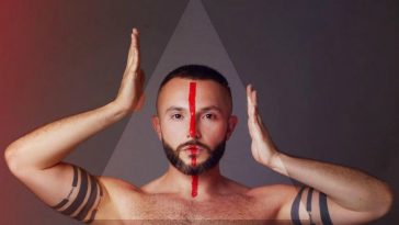 Vasil topless with a red line down the centre of his face, with his hands on either side of his head in a promo shot for his Eurovision song "YOU".