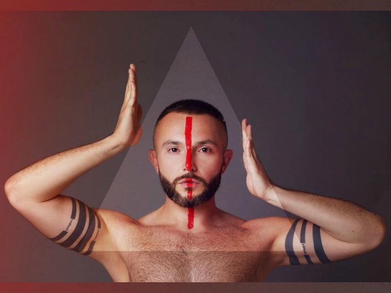 Vasil topless with a red line down the centre of his face, with his hands on either side of his head in a promo shot for his Eurovision song "YOU".