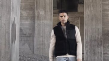 A still from Eric Saade's music video for "Glas" with Eric Saade wearing a black bomber jacket over a cream jumper and light blue jeans standing on gravel amongst grey pillars.