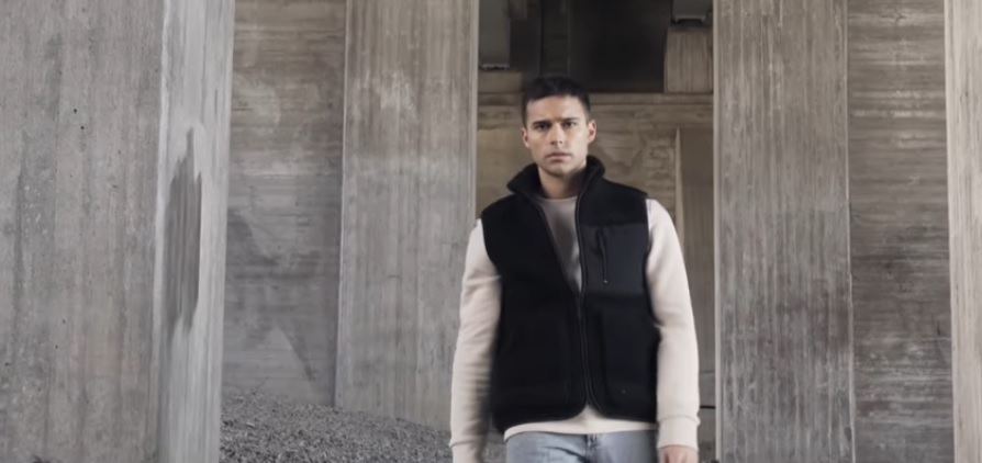 A still from Eric Saade's music video for "Glas" with Eric Saade wearing a black bomber jacket over a cream jumper and light blue jeans standing on gravel amongst grey pillars.