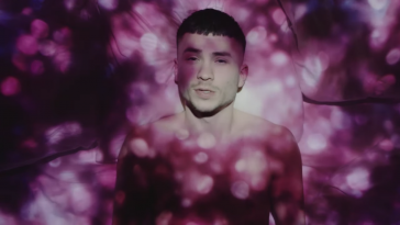 Still from the "Talking In My Sleep" music video which sees Paul Rey lying in a bed with white sheets, topless, as a projector displays pink spotted lights on him and the bed.