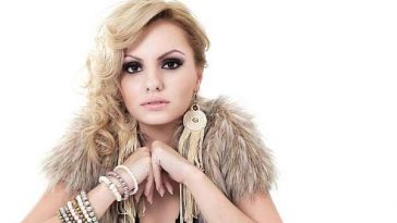 Alexandra Stan promo photo wearing a fluffy shawl around her shoulders with her elbows resting on something in front of her, whilst her blonde hair is wavy, all over one shoulder.