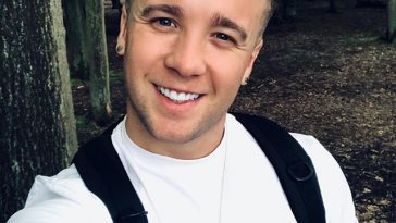 Sam Callahan wearing a white t-shirt and a black backpack in the woods, smiling.