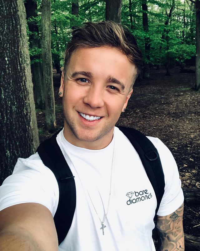 Sam Callahan wearing a white t-shirt and a black backpack in the woods, smiling.