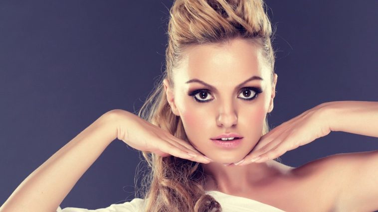 Alexandra Stan framing her face with her hands under her chin and her blonde hair tied up in a ponytail.