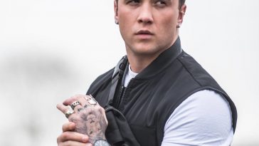 Image of Sam Callahan wearing a white short-sleeved t-shirt with a black bomber jacket, holding his wrist in front of him showing off his tattoo sleeve and his black wrist watch.