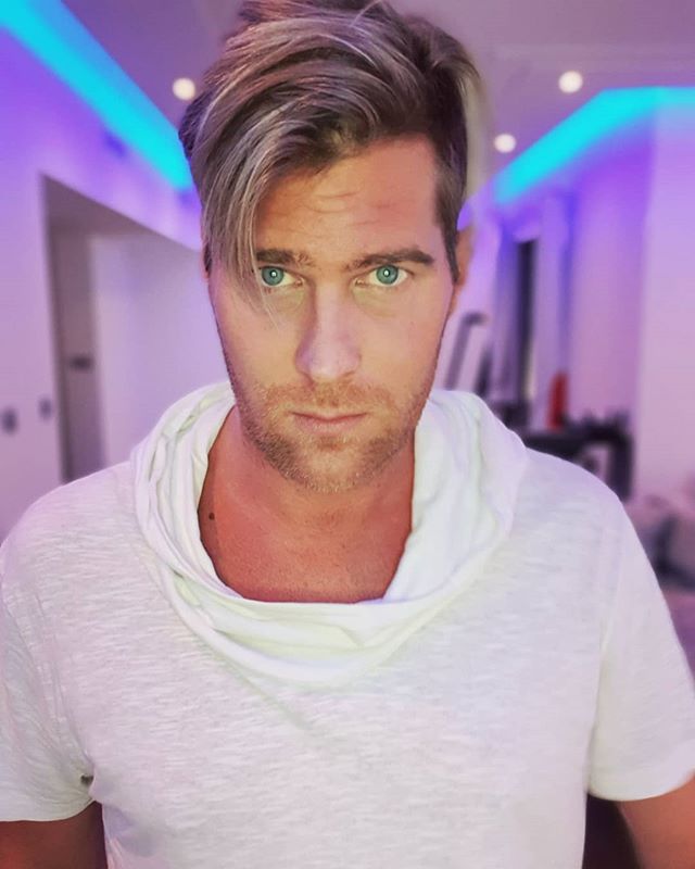 Basshunter taking a selfie wearing a white t-shirt with a bulky collar, and neon blue lights behind him.