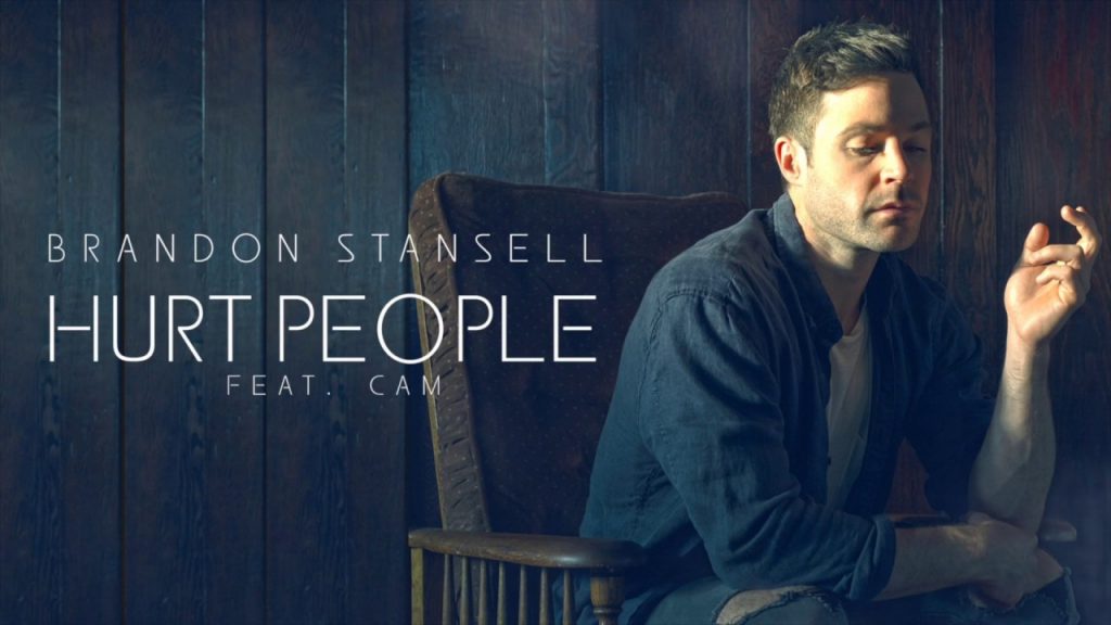 Brandon Stansell, in promotion of new single "Hurt People" sitting in a wooden chair wearing a blue jacket and a white top.