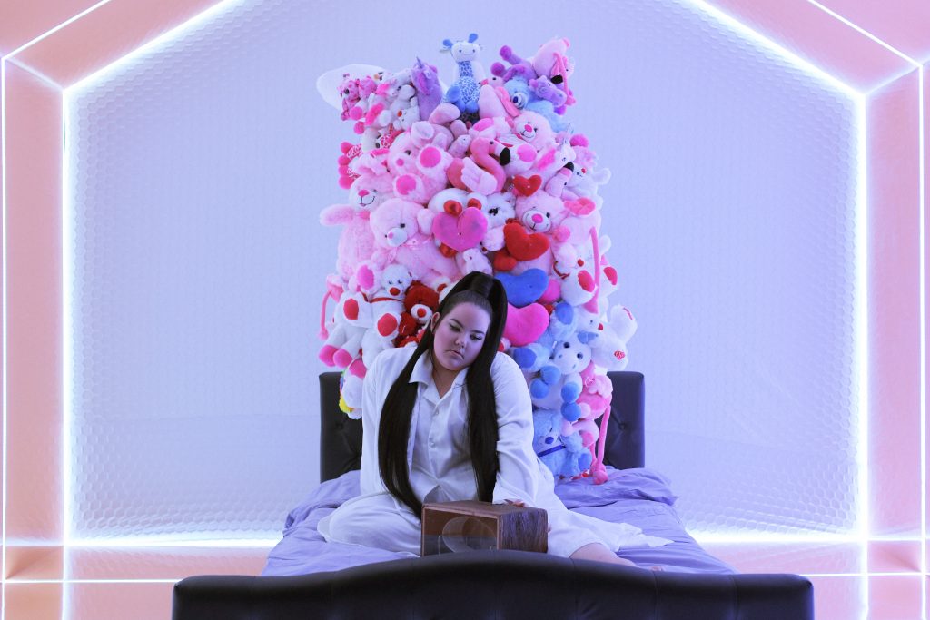 Still from the "Cuckoo" music video which sees Netta sitting on a bed with her music box in front of her in a neon room.