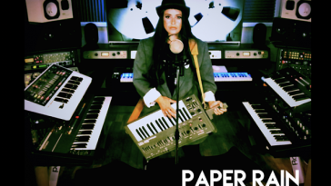 The single cover artwork for "Paper Rain" which sees Lolly in the studio with her keytar and keyboards around her as she sings into a mic.