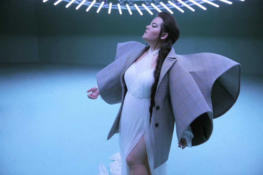 Still from the "Cuckoo" music video which sees Netta in a white dress with an oversized jacket with wing-like sleeves and her hair over one shoulder.