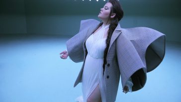 Still from the "Cuckoo" music video which sees Netta in a white dress with an oversized jacket with wing-like sleeves and her hair over one shoulder.