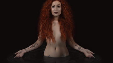 Album artwork for "Confessional" which sees Janet Devlin in a dark pool of water up to her waist, naked and bare with her red auburn hair covering her chest.