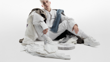 Album artwork for Sillygomania which sees Loïc Nottet sitting with his knees up and his arms on top of his legs, dressed in white with white clothes strewn around him of different colours with a white background.