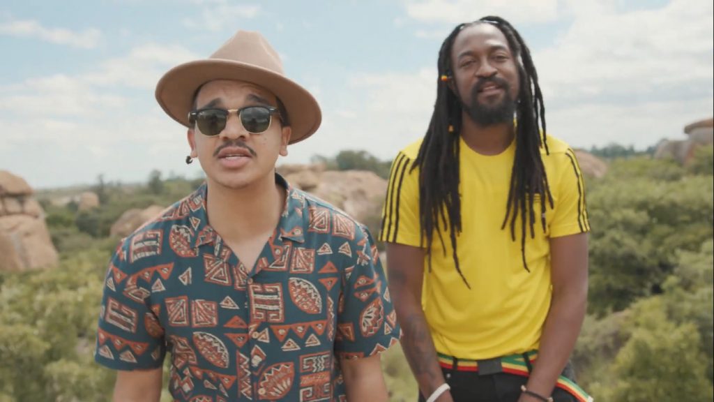 Still from the "Midnight Love" music video with Jindi on the left sporting a sun hat, sunglasses and an Aztec shirt and Smylie on the right wearing a yellow shirt with black dreadlocks down to his chest.