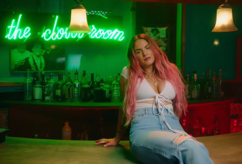 Still from the "What U Need" music video which sees JoJo with pink hair, sitting on a bar top, wearing a white strap bra and high-waist jeans