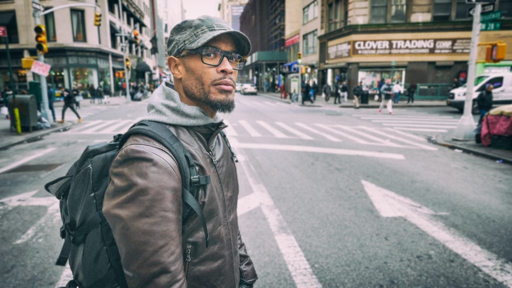 Pete Chatmon wearing a cap and a brown leather coat with a backpack, crossing a city centre street.