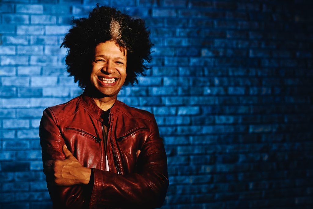Press photo of Marc Lottering laughing at the camera with his arms crossed, donning a deep brown leather jacket.