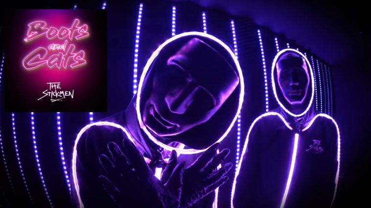 Still from the "Boots And Cats" music video which sees The Stickmen in their signature neon LED stickmen costumes, with the LEDs on the colour purple, there's also LED lights behind them that are the same purple tone, whilst in the top-left corner there is a photo of the single's cover art which has the title in cursive font that's neon pink.