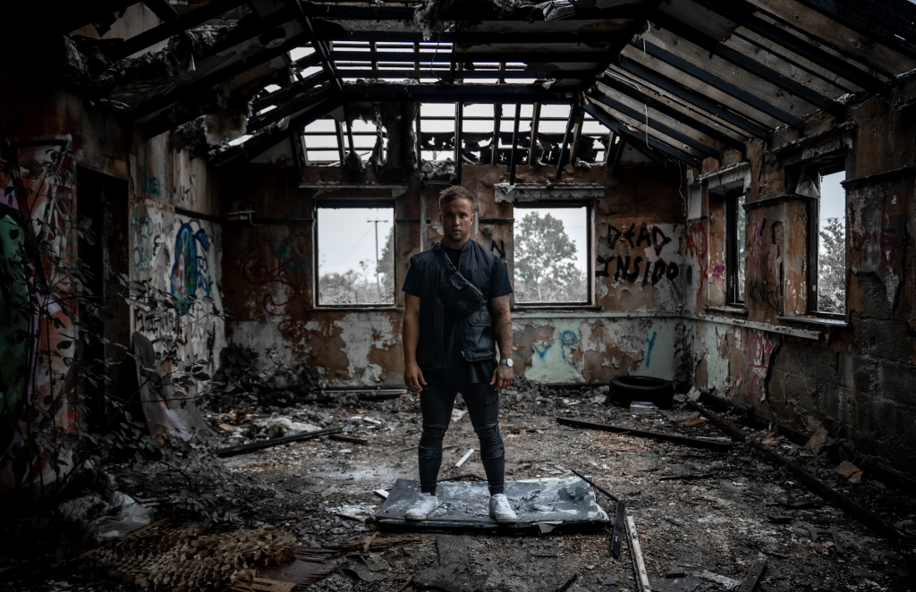 Still from the "Demons" music video which sees Sam Callahan standing on top of a smashed TV within an abandoned building. He is wearing a black t-shirt, a black gilet, black skinny jeans and white trainers.