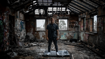Still from the "Demons" music video which sees Sam Callahan standing on top of a smashed TV within an abandoned building. He is wearing a black t-shirt, a black gilet, black skinny jeans and white trainers.