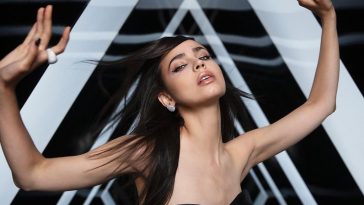 Still from the "Guess I'm A Liar" music video which sees Sofia Carson with her arms up and outstretched with lighted triangles behind.