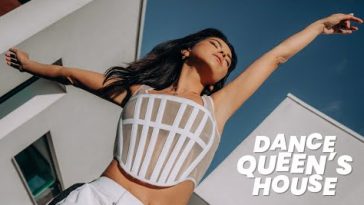 INNA from "Dance Queen's House" day two where she's dancing in front of the buildings with her black hair over one shoulder as she's wearing an armoured-like white top