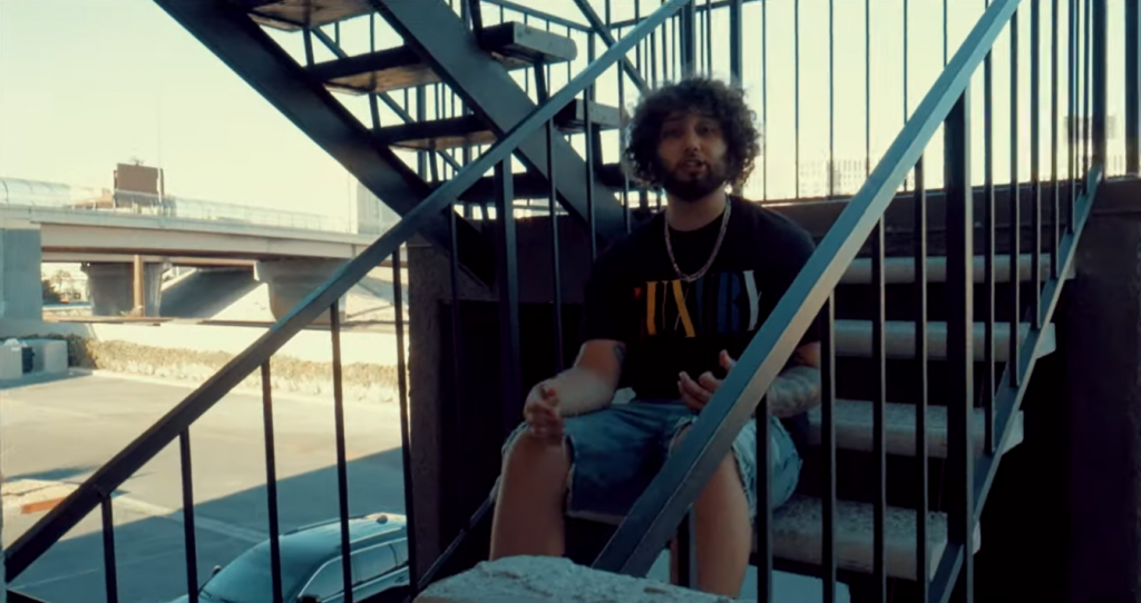 Screenshot from the "Stevie Wonder" music video which sees Vior sitting on a set of stairs wearing jean shorts and a black t-shirt with afro hair and a beard.