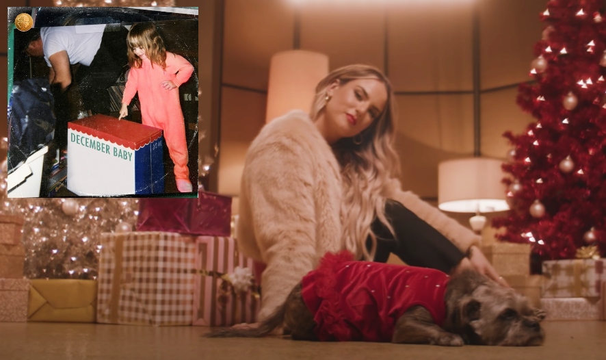 Music video still from "The Christmas Song" video from last year that sees JoJo sitting in front of the camera wearing black jeans and a cream coloured jacket and a red blanket over one leg, sitting on the floor with her blonde hair over one shoulder, looking at the camera, with presents stacked up against her back with a gold Christmas tree behind whilst an image in the top left corner showing the album artwork for "December Baby" which shows JoJo as a child in a pink jumpsuit opening a Christmas box with her dad behind her.