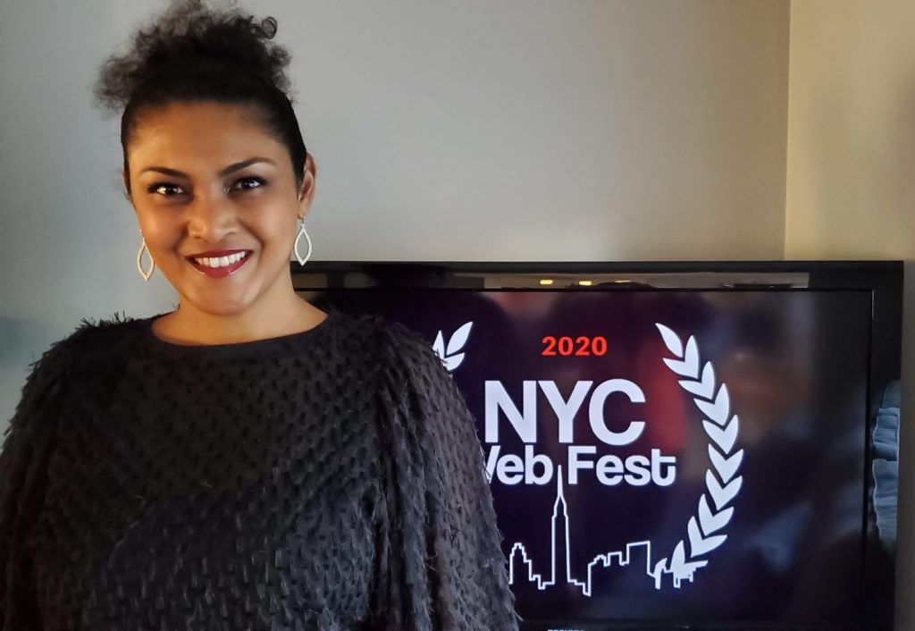 Lauren Atkins wearing a black jumper posing in front of a TV with the 2020 NYC Web Fest logo displayed