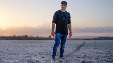 KAIS wearing a black snapback, blue jeans, white trainers and a black t-shirt, standing on a beach.