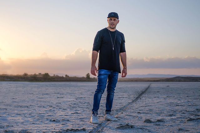 KAIS wearing a black snapback, blue jeans, white trainers and a black t-shirt, standing on a beach.
