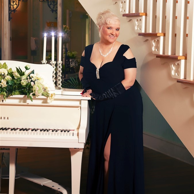 Luanne Hunt stands in the middle of the picture with white hair tied up and in a long black dress. There's a white piano on the left with three burning candles, and a staircase to the right.