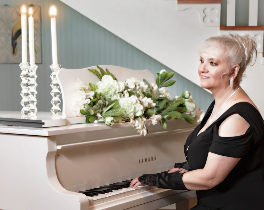 Luanne Hunt sits at a white piano, looking off at the distance with her hands on the piano keys. She's dressed in a black dress and her white hair is tied up.