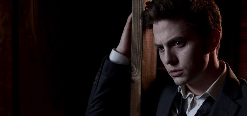 Jackson Rathbone ghostily stares off to the left of the camera in a sultry ghosty way, dressed in a suit with his arm around a wooden pillar.