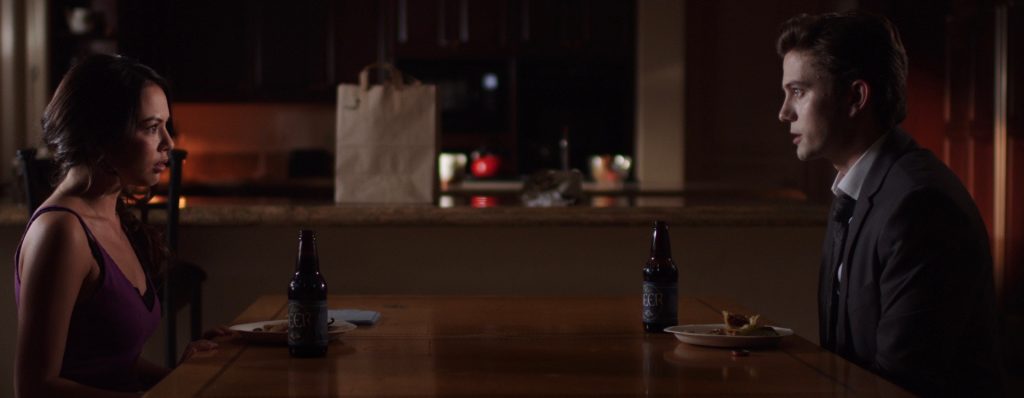 Janel Parrish and Jackson Rathbone sit at a wooden table with food and a beer in front of each of them.