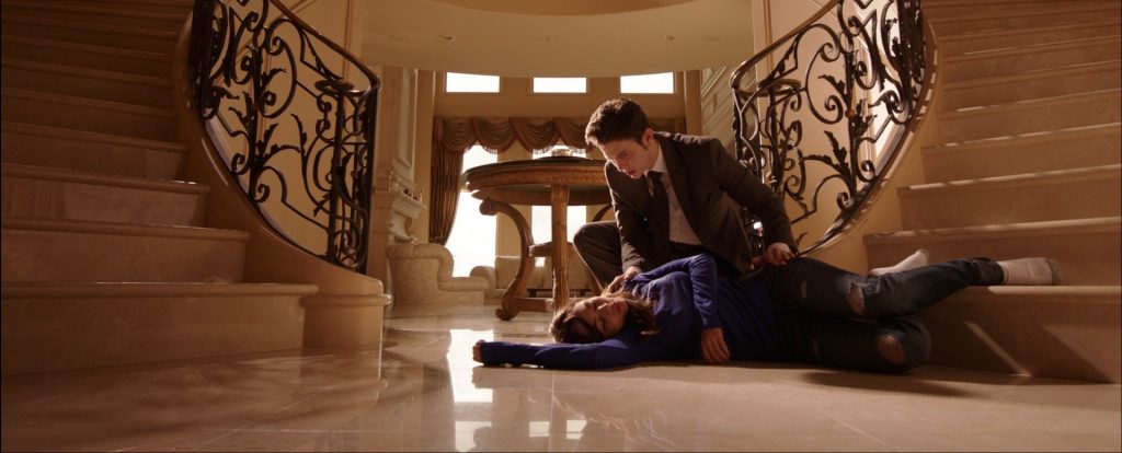 Scene from Until We Meet Again where Janel Parrish is wearing a blue top and black trousers lying at the bottom of a staircase with her eyes shut and Jackson Rathbone is crouched behind her in shock and trying to see if she's okay.